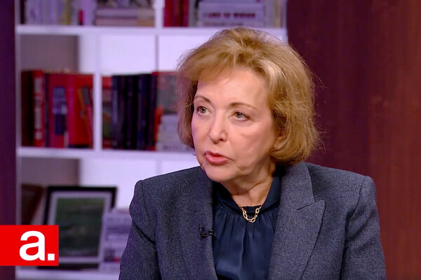 Screenshot of woman on set, seated, with bookshelf in background